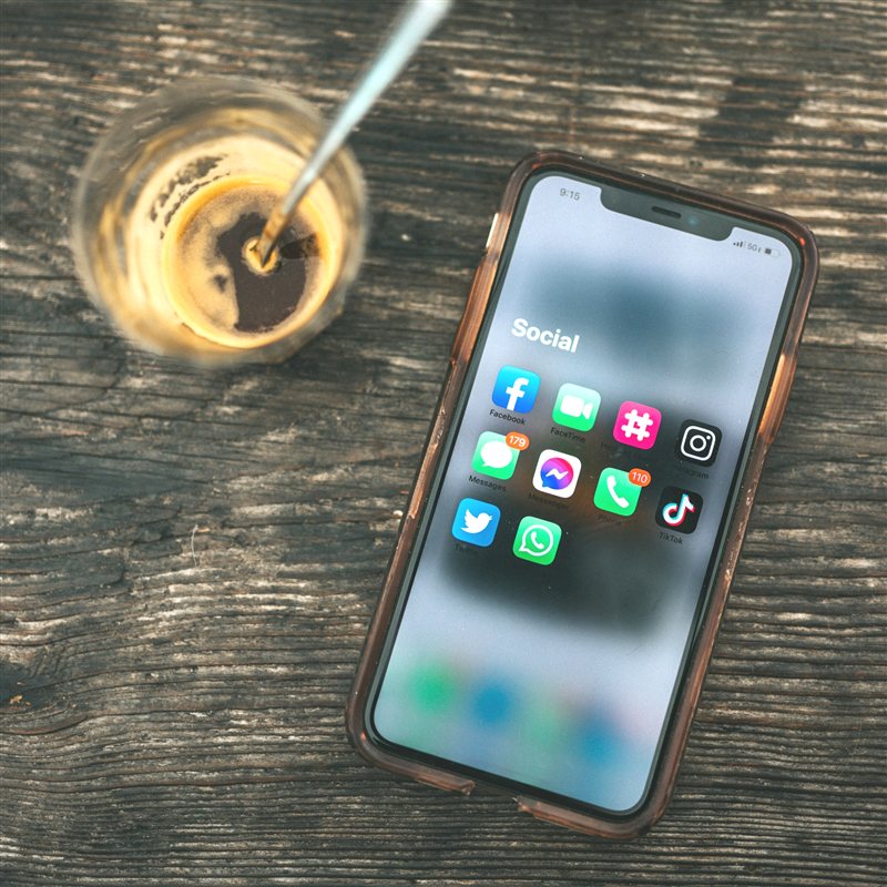 A smartphone lying on a wooden table showing a folder of social and messaging apps, next to a cup of coffee